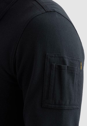 Manga de tela negra texturizada con un bolsillo de parche rectangular y una pequeña etiqueta amarilla de PME Legend en la parte superior del brazo.