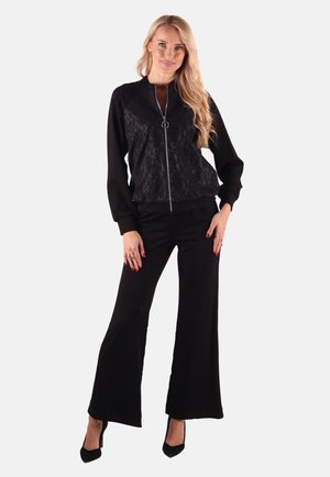 Schwarze Bomberjacke mit Spitzendetails, Reißverschluss und rundem Zipper. Kombiniert mit schwarzen weit geschnittenen Hosen und spitzen schwarzen Absätzen.