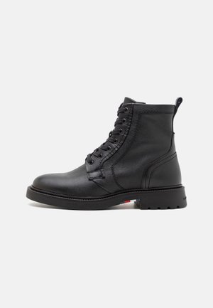 Bota negra de cuero hasta el tobillo con cordones en la parte delantera, lengüeta en el talón y suela resistente con un pequeño detalle rojo, blanco y azul.