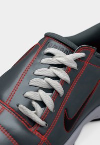 Γκρι αθλητικό παπούτσι με μαύρο λογότυπο Nike, λευκές κορδόνια και έντονες κόκκινες λεπτομέρειες ραφής σε λείο δερμάτινο επάνω μέρος.