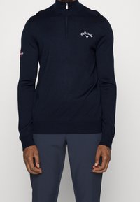 Pull à col zippé en marine foncé, confectionné en tissu doux, avec une coupe ajustée, des poignets côtelés et un logo sur la poitrine et la manche.