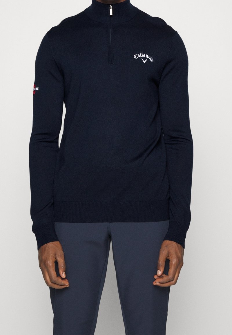 Pull à col zippé en marine foncé, confectionné en tissu doux, avec une coupe ajustée, des poignets côtelés et un logo sur la poitrine et la manche.