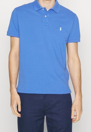Mann trägt ein blaues Polo mit kurzen Ärmeln und dunkelblaue Hose und steht vor einem einfarbigen Hintergrund.