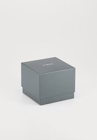 Cutie din carton gri, cu o formă pătrată, având un capac plat și un model subtil embosat. Pe față este inscripționat cuvântul "TIMEX" în alb.