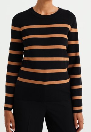 Personne portant un pull noir à manches longues avec des rayures horizontales marron et un pantalon noir, debout devant un fond blanc uni.