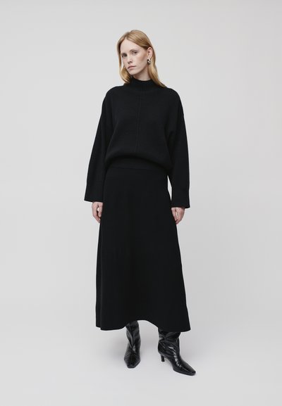 Schwarzer gestrickter Pullover mit hohem Kragen und überschnittenen Schultern, kombiniert mit einem passenden langen Rock. Das Model trägt schwarze Stiefeletten mit Absatz.