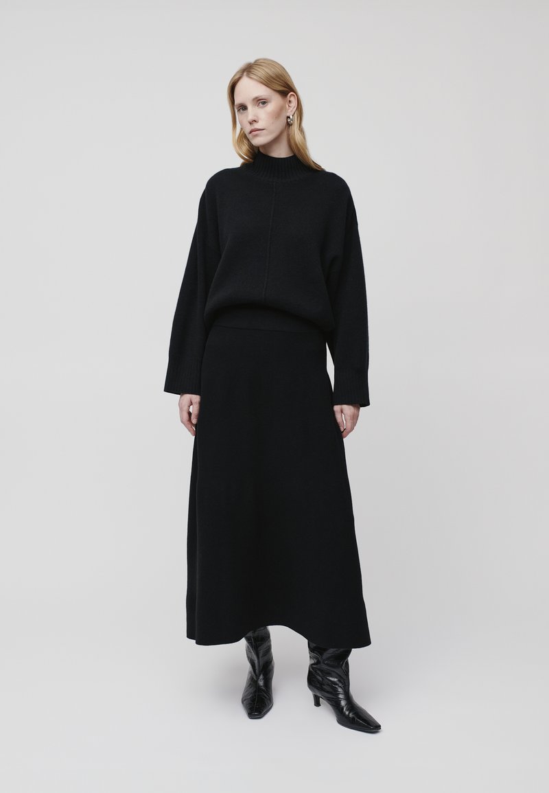 Maglione nero a maglia con collo alto e spalle scese, abbinato a una lunga gonna coordinata. La modella indossa stivaletti neri con tacco.