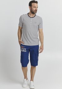 Camisa henley de manga corta a rayas en blanco y azul marino; combinada con pantalones cortos de cargo azules con bolsillos y detalles impresos. Zapatillas blancas.
