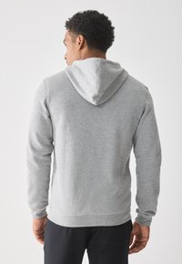 Mann mit kurzen lockigen Haaren, der einen hellgrauen Kapuzenpullover und schwarze Hosen trägt und mit dem Rücken zu einem schlichten weißen Hintergrund steht.