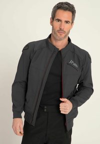 Giacca uomo grigia scura con chiusura lampo frontale, dettagli rossi e spalline. Presenta un logo sul petto e diverse tasche.