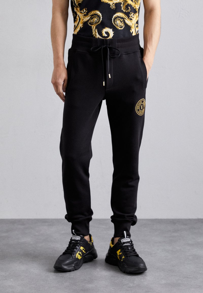 Pantalones de chándal negros con cintura ajustable, que presentan un parche de logo redondo. El modelo lleva zapatillas negras y amarillas con un diseño texturizado.