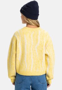 Geel gebreid trui met een losse pasvorm en ribbelzoom, versierd met witte golvende lijnen. Gepaard met een marineblauwe gebreide beanie en blauwe spijkerbroek.