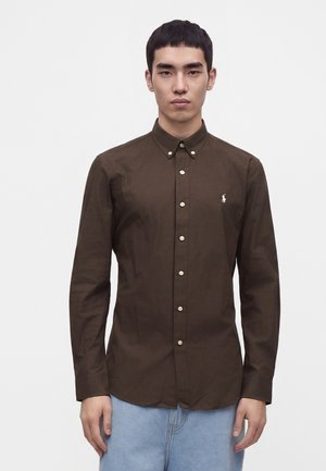 Jeune homme aux cheveux courts et noirs portant une chemise marron foncé boutonnée et un jean bleu clair, debout devant un fond blanc uni.