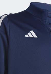 Marineblaue Sportjacke mit hohem Kragen, Reißverschluss und weißen Streifen. Mit einem Logo auf der linken Seite in weißer Stickerei.