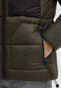 Dunjacka i olivgrön med svart bröstpanel; har en zip-ficka och justerbar dragsko i nederkant. Texturerat, lätt material.