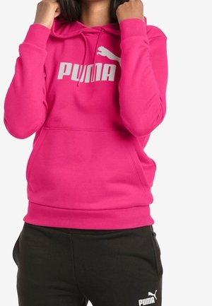 Osoba ubrana w jaskraworóżową bluzę z kapturem marki Puma z logo na piersi oraz czarne spodnie Puma z logo na udzie, ściągająca sznurki kaptura.