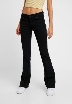 Femme portant un jean flare noir taille haute, des baskets blanches et un crop top sans manches jaune à rayures, sur un fond uni.