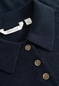 Granatowy dzianinowy polo z kołnierzykiem, posiadające trzy beżowe guziki. Metka z napisem "NOELLA", rozmiar S/M, z dołączoną metką pielęgnacyjną.