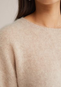 Gai bēša adīta džemperis ar apaļu kakla izgriezumu. Mīksta, pūkaina tekstūra ar smalku melnbaltu rakstu un ribota kakla izgriezuma detaļa.