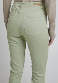 Pantalon en denim vert clair, avec une taille haute, deux poches arrière et une coupe droite avec une couture minimale visible.