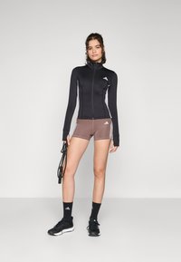 Svart zip-up treningsjakke med lange ermer, kombinert med brune shorts med hvite striper. Tilbehør inkluderer svarte joggesko og sokker.