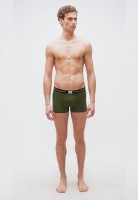 Άνδρας χωρίς πουκάμισο με κατσαρά μαλλιά που φοράει εσώρουχα Polo Ralph Lauren χακί, όρθιος ξυπόλυτος μπροστά σε απλό ανοιχτό φόντο.