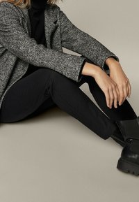 Grijze textuur blazer over een zwarte turtleneck, gecombineerd met strakke zwarte broek en stevige zwarte enkelboots, zittend tegen een neutrale achtergrond.