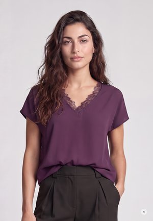Frau mit langen, welligen Haaren, die eine violette Spitzen-V-Ausschnitt-Bluse und schwarze hoch taillierte, plissierte Hose vor einem schlichten Hintergrund trägt.