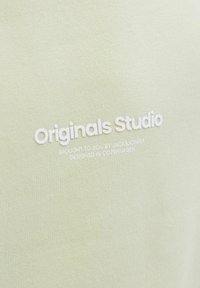 Világoszöld pulóver, texturált anyagból készült, fehér nyomott logóval, amelyen az "Originals Studio" felirat, és további szöveg található alul.