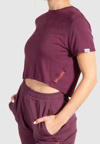 T-shirt bordeaux coupé, à manches courtes, en tissu doux. Présente un petit logo sur l'ourlet et est associé à un pantalon de jogging assorti.