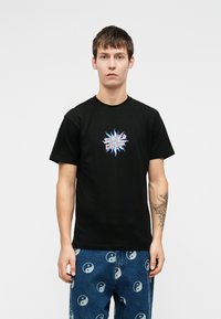 Noor mees kannab musta T-särki, millel on värviline "Santa Cruz" tekst ja päikese graafika, koos siniste teksadega, millel on valged yin-yang sümbolid.