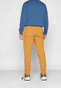 Persona de espaldas, vistiendo sudadera azul y pantalones deportivos amarillo mostaza, con zapatillas deportivas. El fondo es de un color claro y liso.