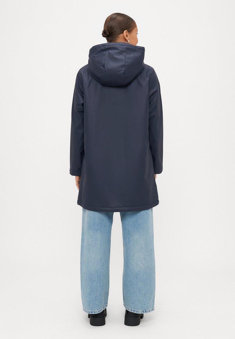 Manteau imperméable bleu marine avec capuche, design long jusqu'aux cuisses et manches longues, associé à un jean large bleu clair et des bottes noires.