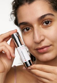 Vit serumflaska med en glänsande silverkapsyl, märkt "Visible Difference Good Morning Primer," som hålls jämte ett ansikte med naturlig hudton.