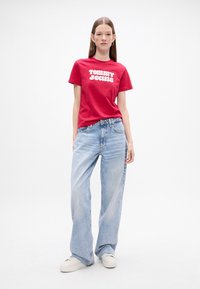 Punane puuvillane T-särk, millel on valge "TOMMY JEANS" trükk, koos helesiniste laia säärega teksade ja valgete tossudega.