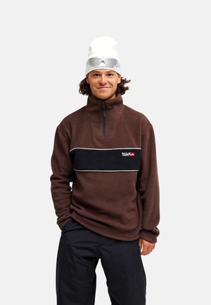 Jeune homme portant un pull polaire marron, un pantalon noir, un bonnet blanc avec des lunettes de soleil posées dessus, debout devant un fond blanc.