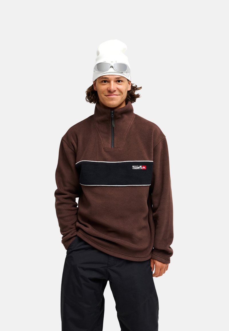 Jeune homme portant un pull polaire marron, un pantalon noir, un bonnet blanc avec des lunettes de soleil posées dessus, debout devant un fond blanc.