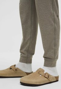 Tan ruskindeklædte træsko med lukket tå, justerbar rem og korksål, parret med lysegrå sweatpants, hvide sokker og kontrasterende syninger.