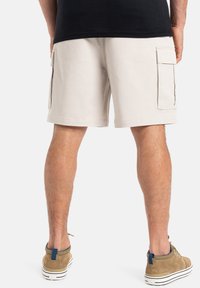 Pantalones cortos cargo beige con dos bolsillos laterales, tejido de algodón, corte recto y acabado en el dobladillo. Combinados con zapatillas de ante marrón.