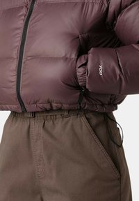 Giacca imbottita viola con chiusura frontale con zip e polsini elastici, abbinata a pantaloni marroni con coulisse e tasche laterali.