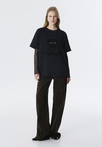Zwarte T-shirt met "BOLD" tekst en een berenafbeelding, gecombineerd met bruin luipaardprint wijde broek. Beide hebben een ontspannen silhouet.