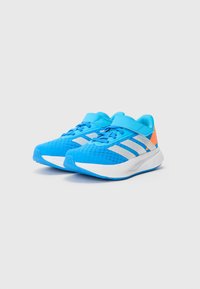 Zapatillas deportivas azules con parte superior de malla, suela intermedia blanca, rayas grises y un acento naranja en el talón. Punta redondeada y cierre con cordones.