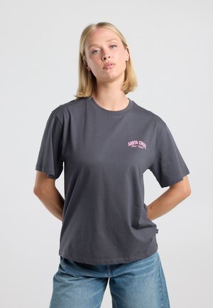 Jonge vrouw met kort blond haar draagt een donkergrijs T-shirt met roze "Santa Cruz West Coast" tekst en blauwe spijkerbroek, staand voor een effen achtergrond.