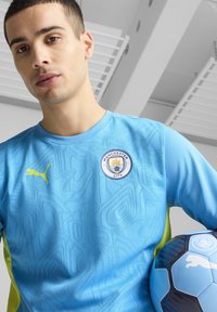Gaiši zila futbola krekls ar teksturētu rakstu, dzeltens Puma logotips un Mančestras City emblema. Tur rokās atbilstoša zila futbola bumba.