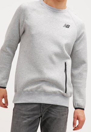 Mężczyzna noszący jasnoszary sweatshirt z okrągłym dekoltem, z czarną kieszenią na zamek po boku oraz czarnym logo na piersi.