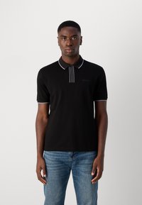 Polo shirt noir avec un col présentant une bordure blanche et des rayures verticales sur le plastron. Fabriqué en tissu lisse, associé à un jean bleu clair.