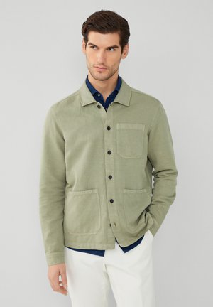 Lichtgroene jas van katoen, met een knoopsluiting aan de voorkant, twee voorzakken en een eenvoudige kraag, gedragen over een marineblauw shirt.