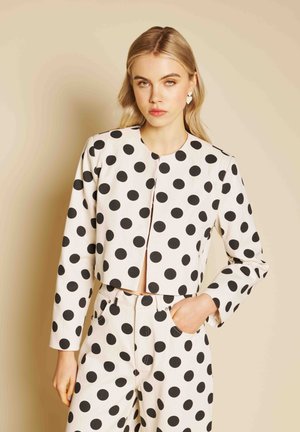 Femme aux cheveux blonds portant une veste blanche courte et un pantalon taille haute à gros pois noirs, debout devant un fond beige.