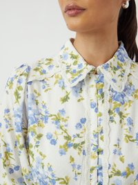 Blouse blanche à motifs floraux bleus et verts, avec un col festonné et une délicate bordure en dentelle. Présente des boutons sur le devant.