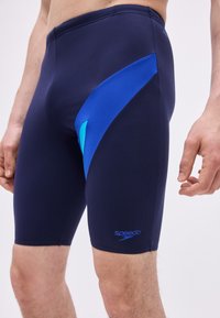 Άνδρας που φοράει μαγιό jammers σε ναυτικό μπλε με μπλε λεπτομέρειες και λογότυπο Speedo στο δεξί μηρό, όρθιος με τα χέρια χαλαρά στα πλάγια.